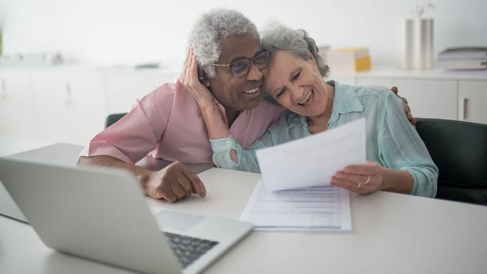 Two older women reading a document together at a table beside a laptop