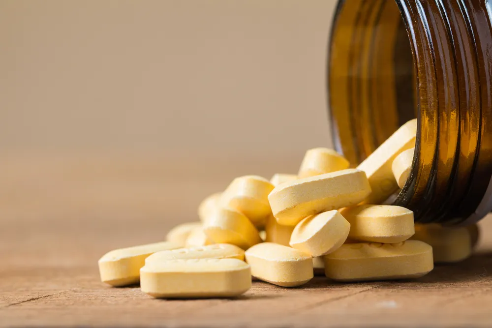 An amber bottle tipped over with pale yellow supplement tablets spilling onto a wooden table