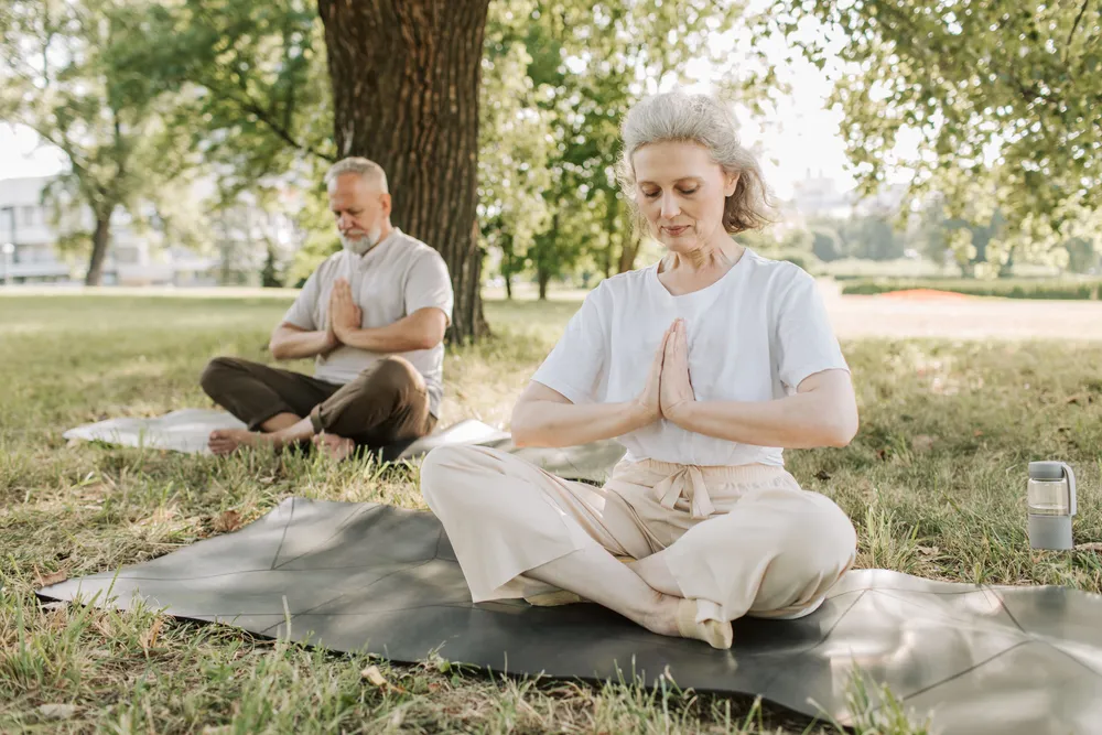 An older couple sitting cross-legged on mats doing yoga in a park